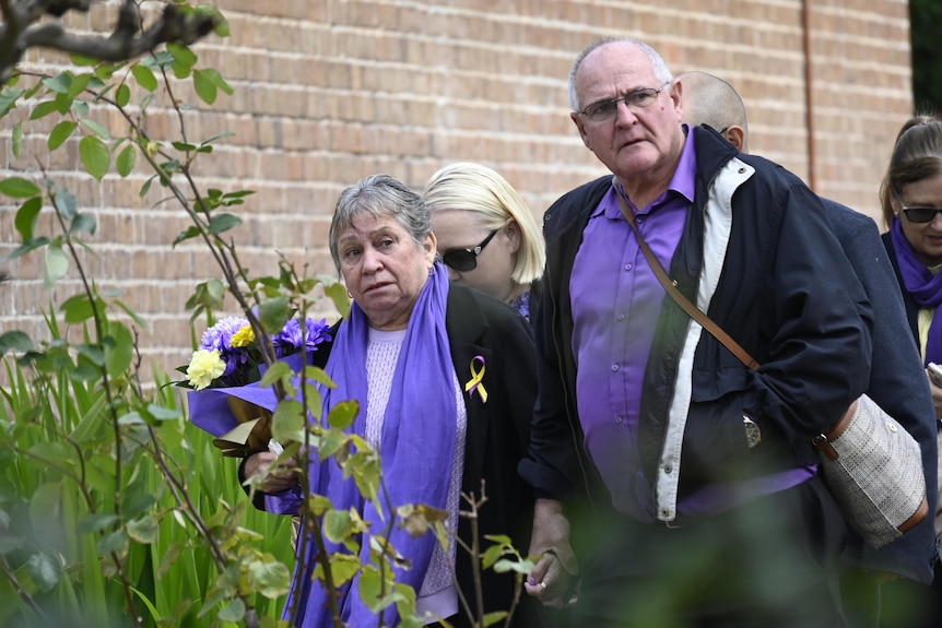 And older woman and man, wearing purple and holding flowers, walk through a garden outside a brick building, looking upset.