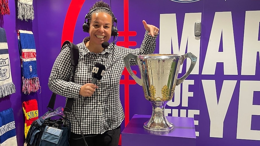woman with headphones on holding microphone pointing downward at large golden cup trophy on a purple stand