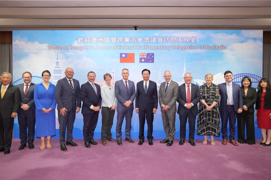 A group of Australian and Taiwanese delegates pose for a photo in front of a large banner with images of both flags.  