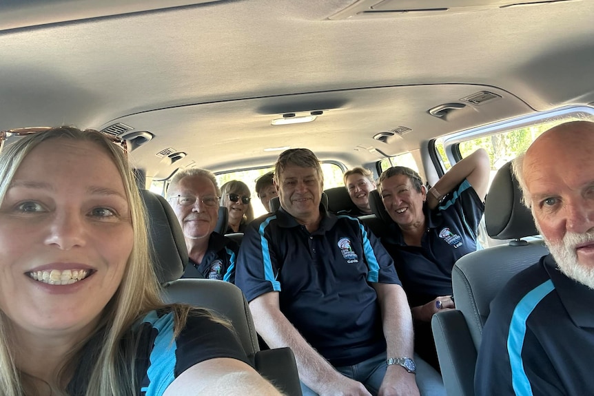 A blonde woman smiles in selfie with a van full of older adults wearing navy blue shirts with blue detail. They all smile.