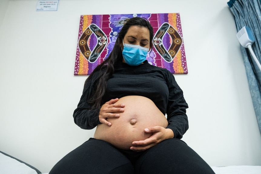 A deeply pregnant woman holds her belly in front of an Indigenous artwork