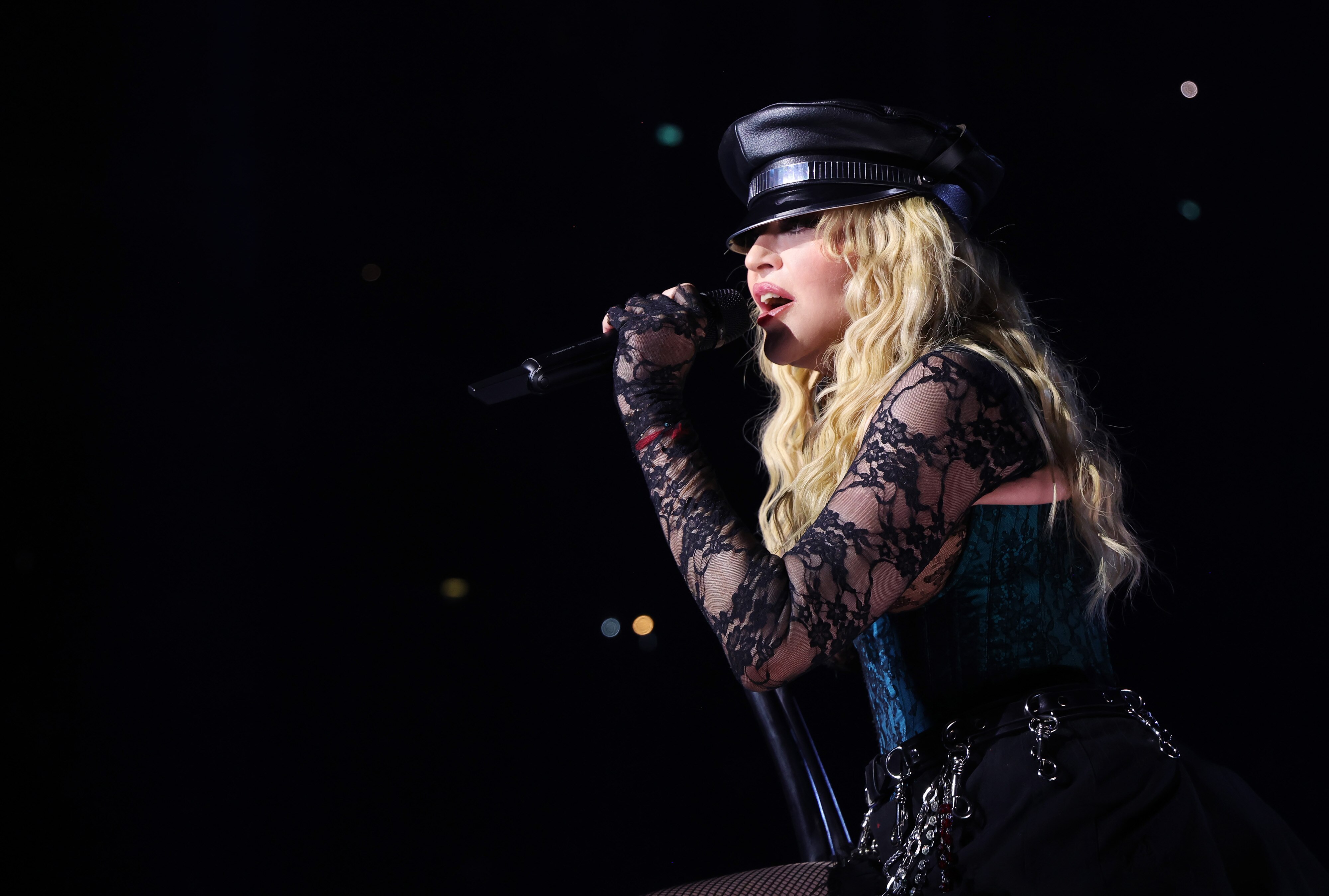 A close up of Madonna, wearing a hat and lacey sleeves, singing into a microphone during a concert.