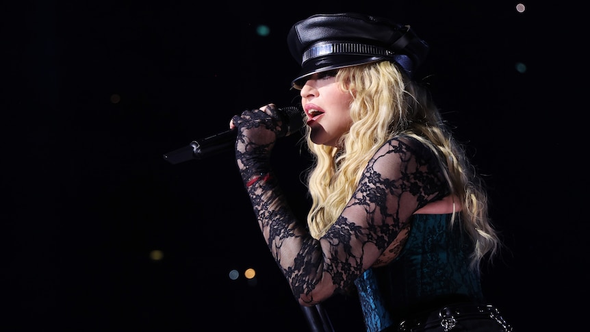 A close up of Madonna, wearing a hat and lacey sleeves, singing into a microphone during a concert.