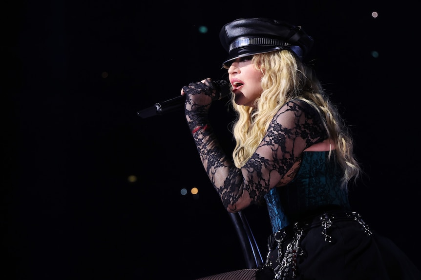 A close up of Madonna, wearing a hat and lacey sleeves, singing into a microphone during a concert.