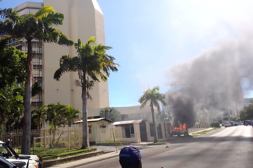 A vehicle on fire in the street outside a building. 