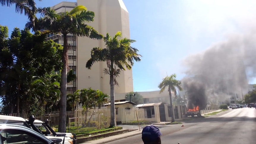 A vehicle on fire in the street outside a building. 