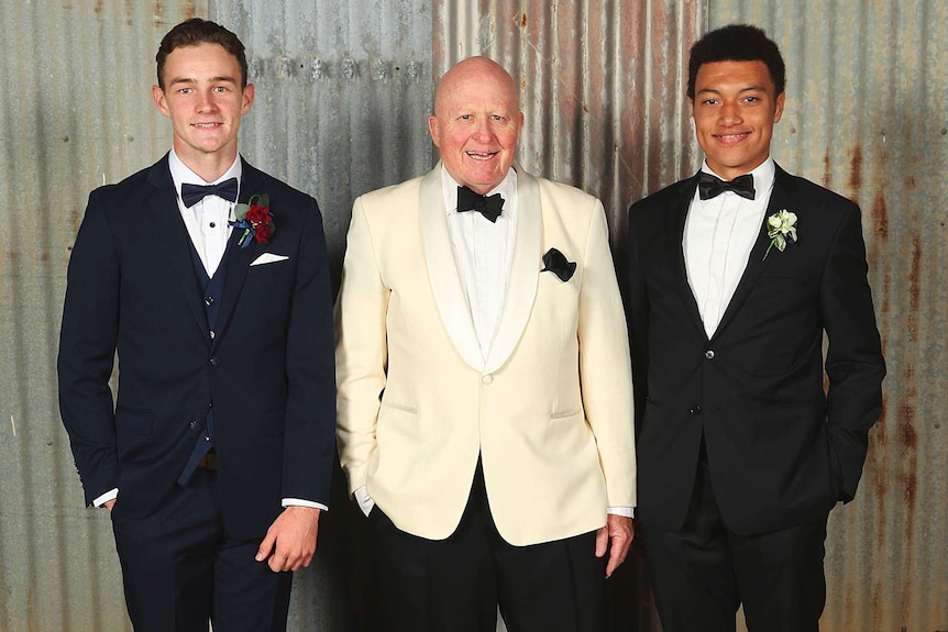 Man with white tuxedo and black bow tie standing between two young men in formal attire.