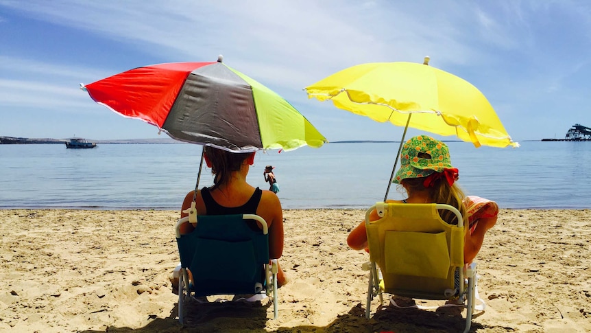 Two children sit in chair under the cover to umbrellas at the beach.