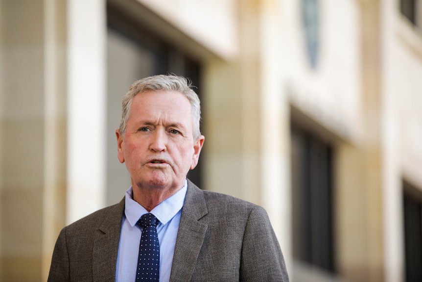 A head and shoulders shot of Shane Love who wears a grey jacket and blue shirt and tie