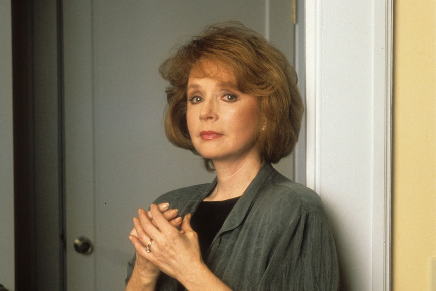 Piper Laurie with red hair looking at the camera and leaning against the wall with her hands together