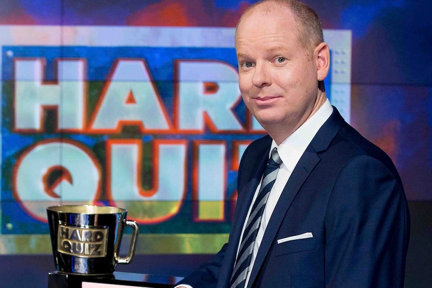 Tom Gleeson poses next to a trophy with the words 'Hard Quiz' visible in the background.