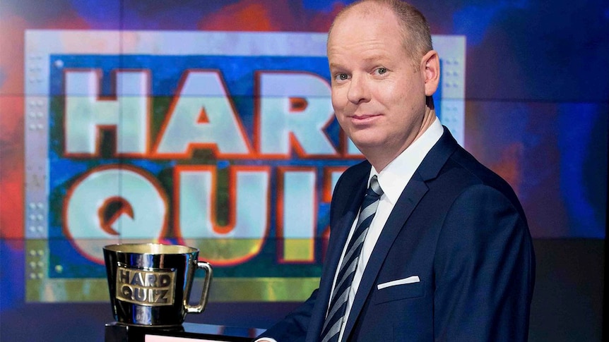 Tom Gleeson poses next to a trophy with the words 'Hard Quiz' visible in the background.