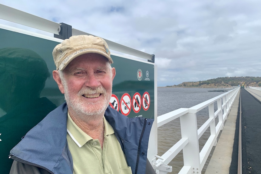 A man stands on a bridge with a sign behind him that shows which activities are prohibited on Granite Island