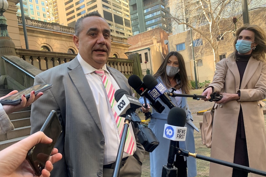 A man wearing a suit stands in front of microphones surrounded by a number of journalists.