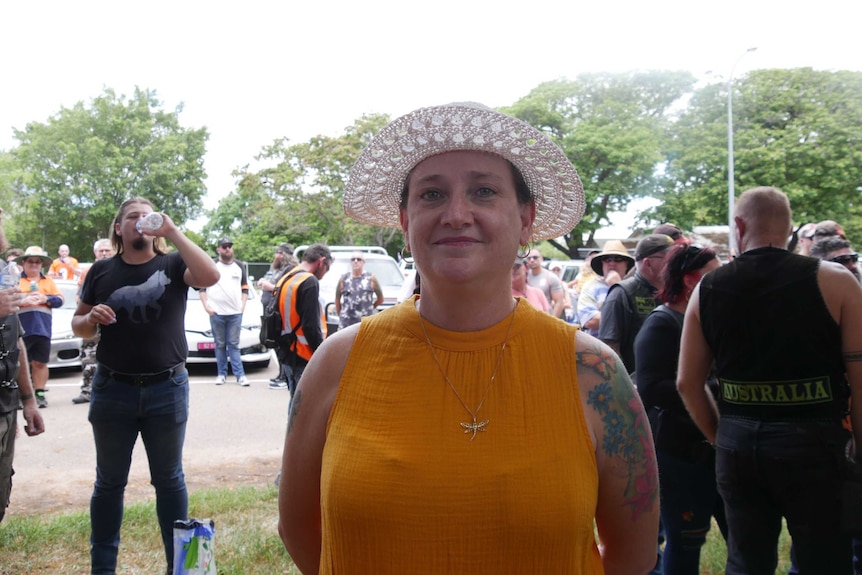 Julianne Wood looks to the camera with a small smile. She wears an orange top and a white hat in front of a crowd.