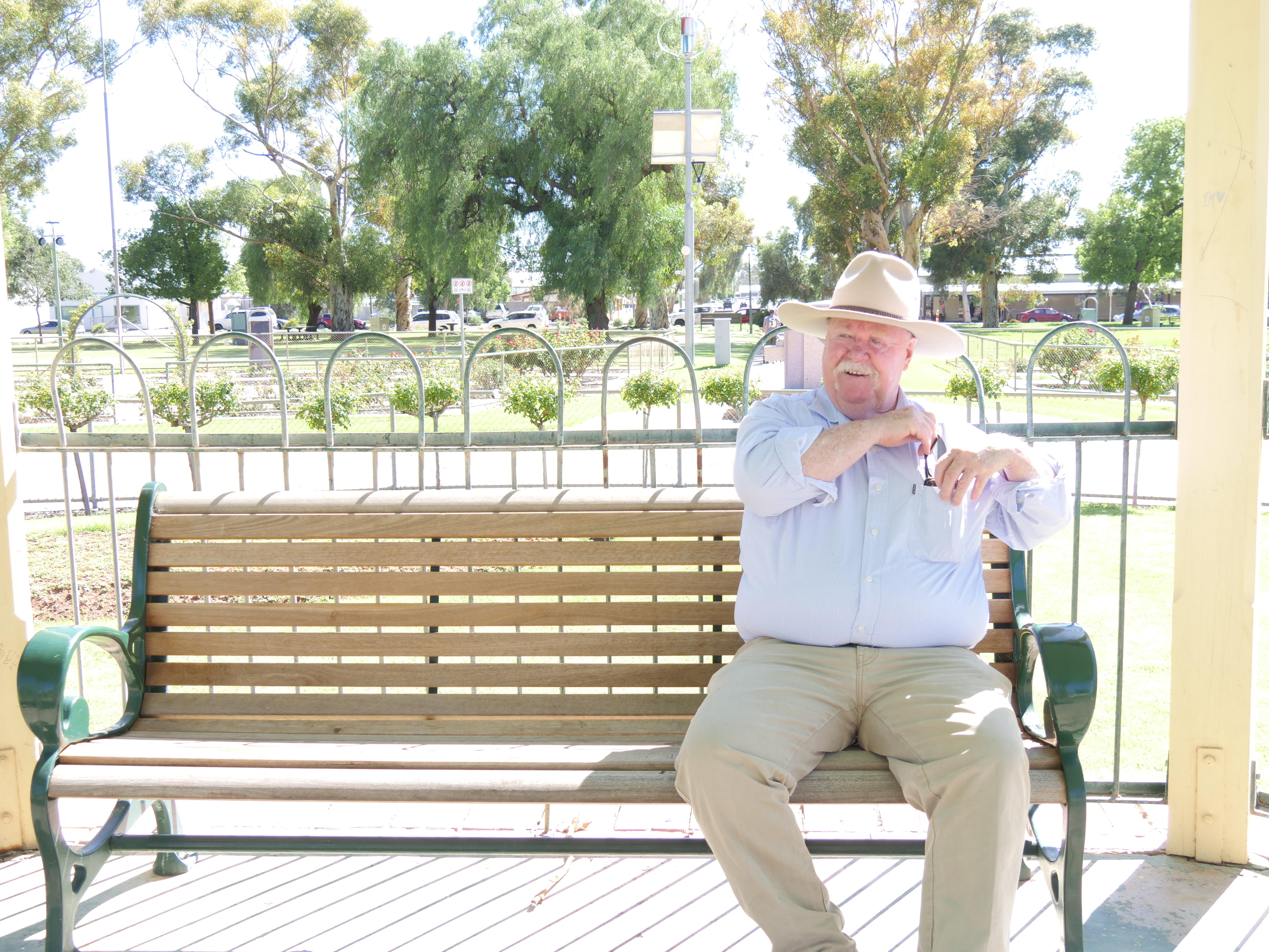 A Caucasian man in a hat sits on a park bench, putting something in the pocketof his light shirt, khaki pants, greenery behind.
