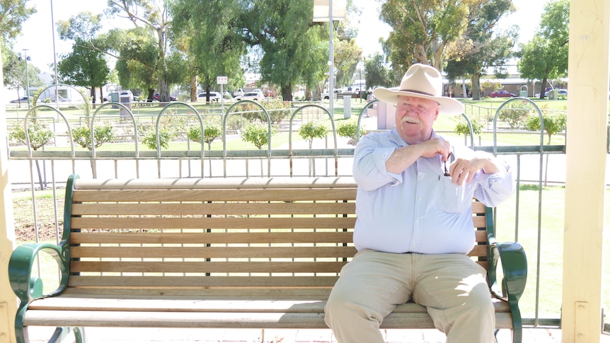 A Caucasian man in a hat sits on a park bench, putting something in the pocketof his light shirt, khaki pants, greenery behind.