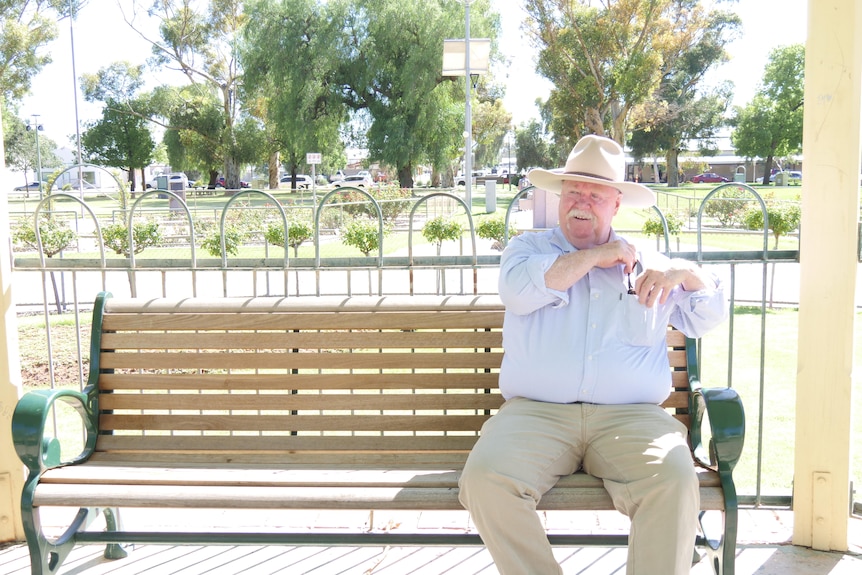 A Caucasian man in a hat sits on a park bench, putting something in the pocketof his light shirt, khaki pants, greenery behind.