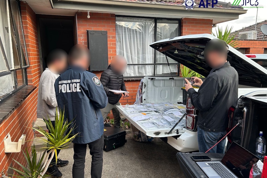 People, including police, gather around an open car tray viewing blurred items outside a house.