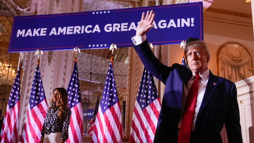 A man in a suit waves his right hand as he stands before a large banner that says: "Make America Great Again".