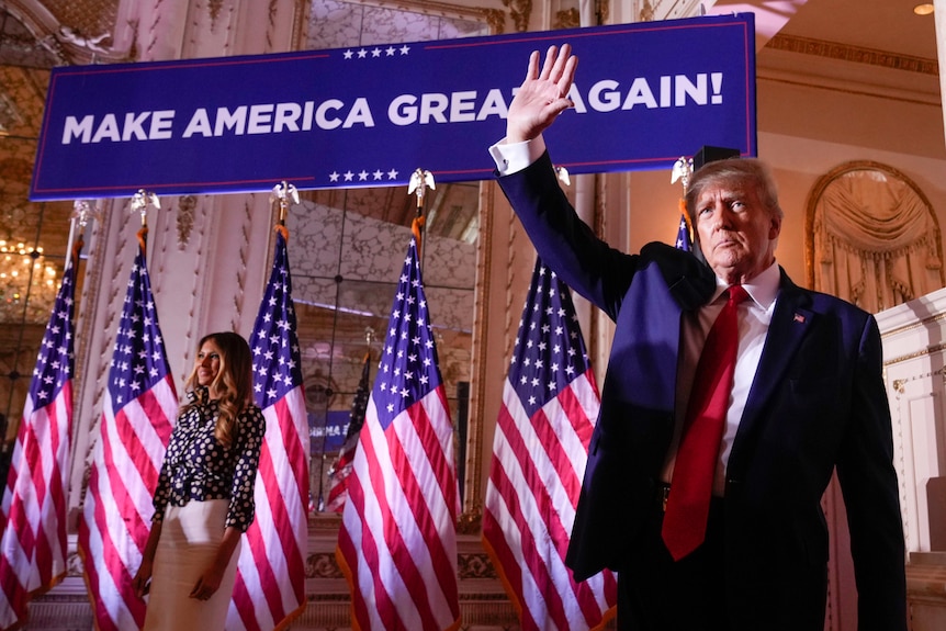 A man in a suit waves his right hand as he stands before a large banner that says: "Make America Great Again".