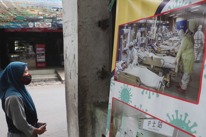 A woman wearing a hijab and face mask looks at a coronavirus-themed banner in Jakarta.