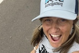 A selfie of a smiling woman with her hair in plaits, a light blue cap and a grey singlettop. 