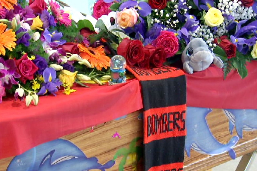 Brightly-coloured flowers, a toy dolphin and an Essendon "Bombers" scarf top the wooden coffin of Noeline Dalzell.