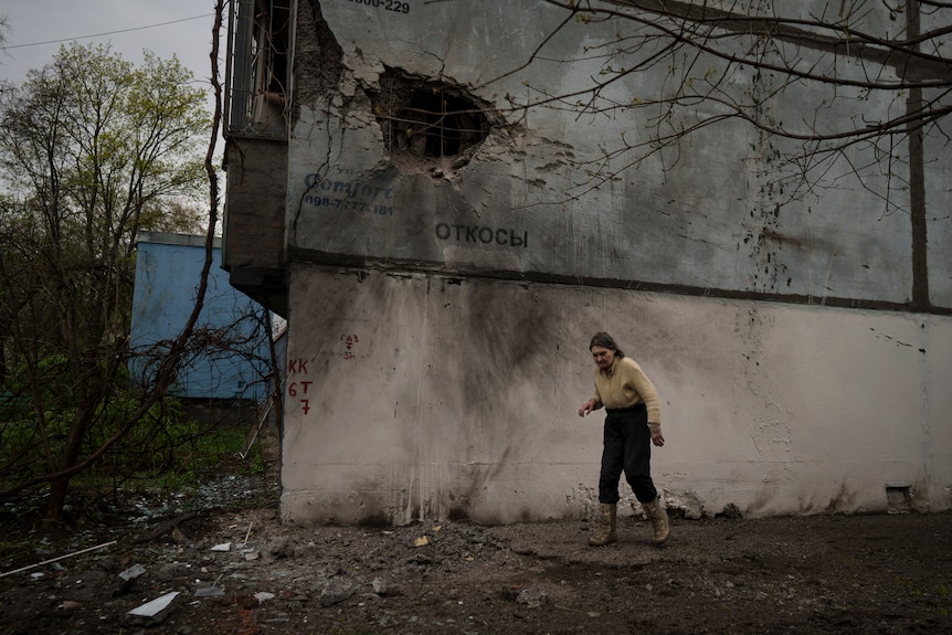 A woman walks past a damaged building with a huge hole blasted into the side 