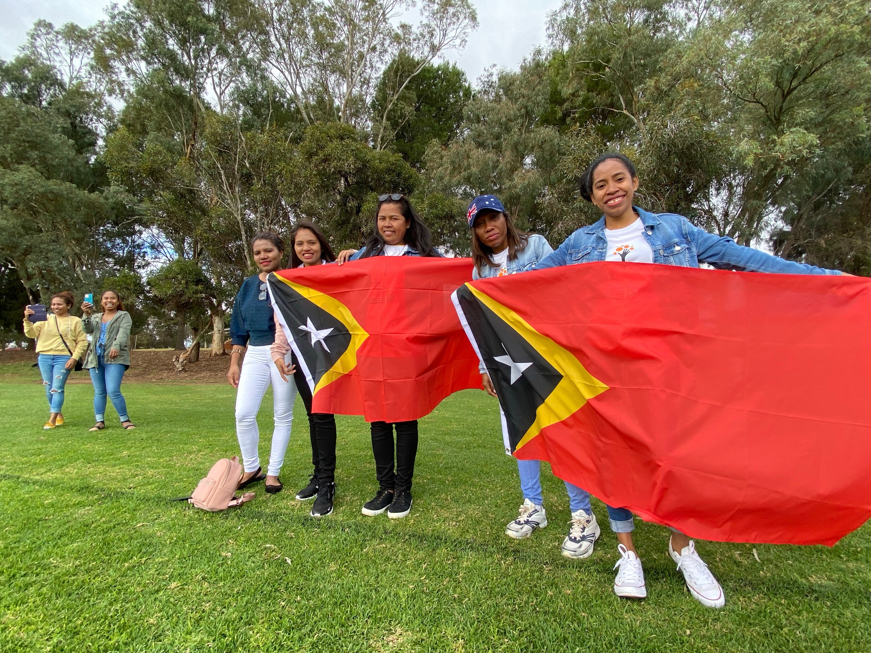 Five women hold up two East Timorese flags as they cheer their team mates