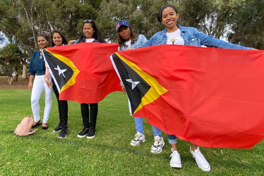 Five women hold up two East Timorese flags as they cheer their team mates