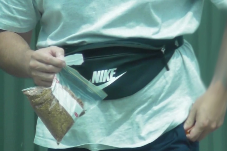 A man with a bum bag holds a bag of tobacco and a rolling paper.