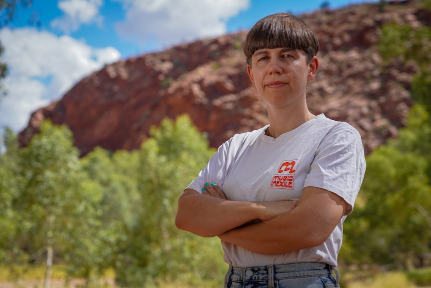 A woman looking serious and standing with her arms crossed, outside in front of red rocks and greenery.