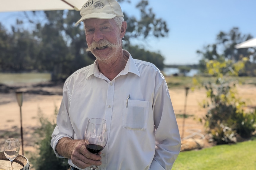 Eric, a fair-skinned man, with white hair and moustache, smiles as he holds a glass of red wine under a shade umbrella.