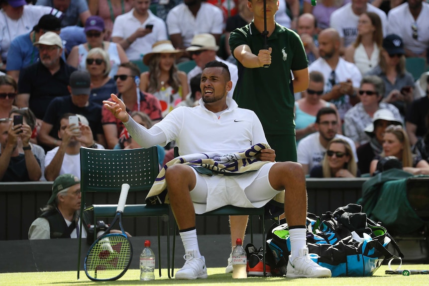 Nick Kyrgios sits on a chair with his legs wide apart and gestures with one hand, looking frustrated.