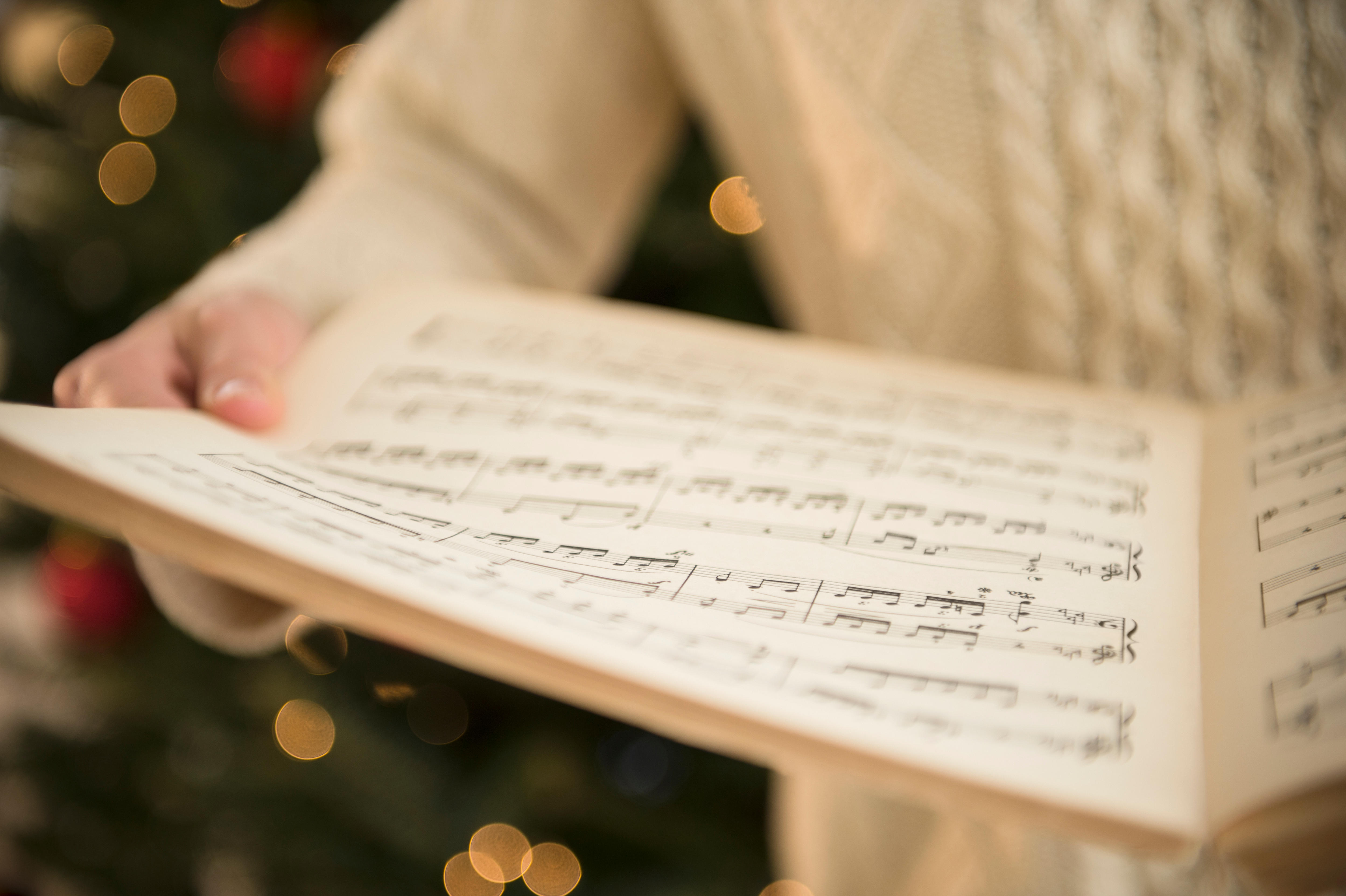 A woman's hand in a white sweater holding sheet music with Christmas lights in the background