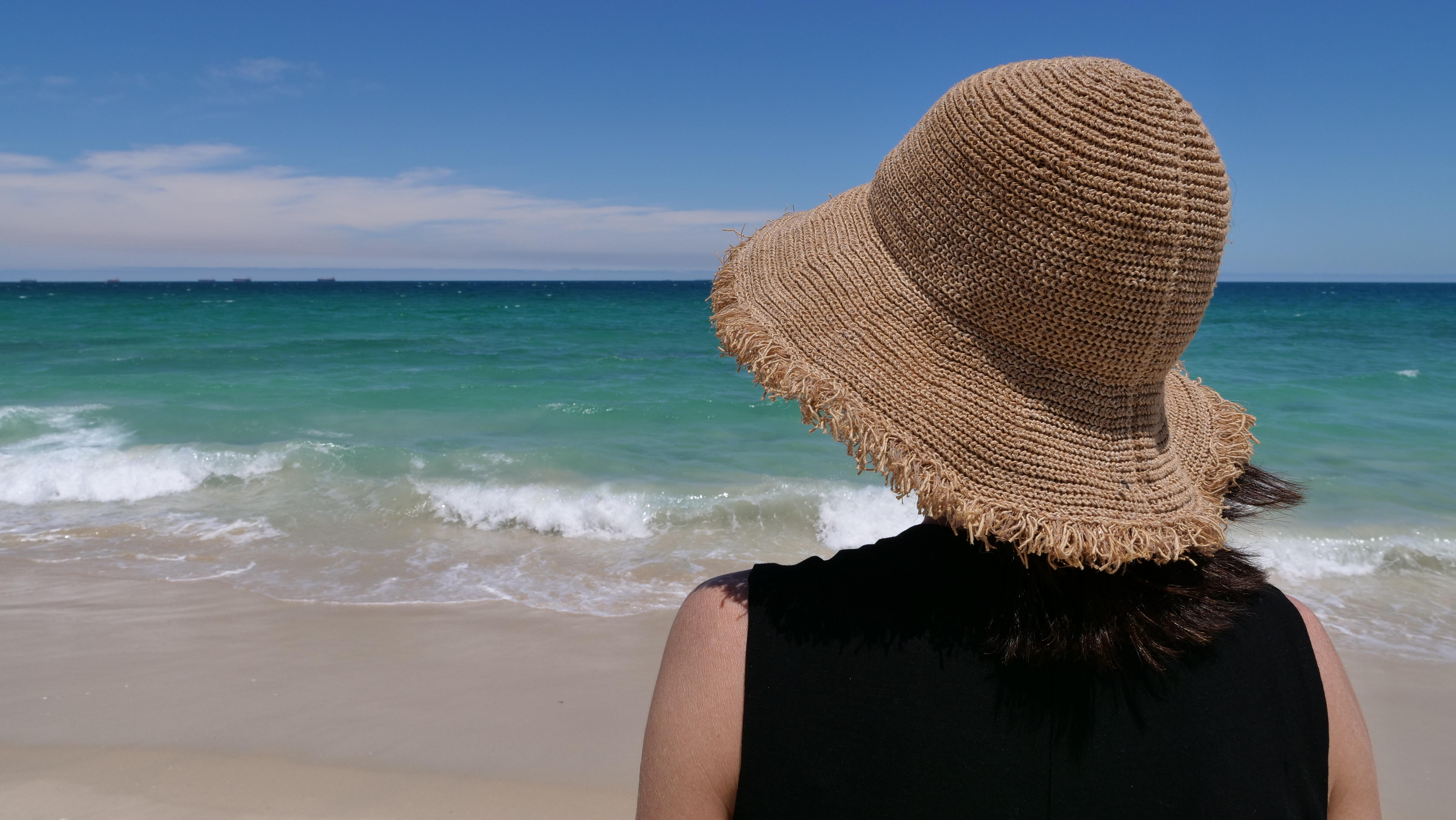 A picture of a woman from behind, wearing a strawhat at the beach.