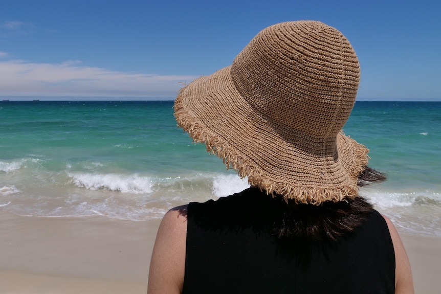 A picture of a woman from behind, wearing a straw hat at the beach.