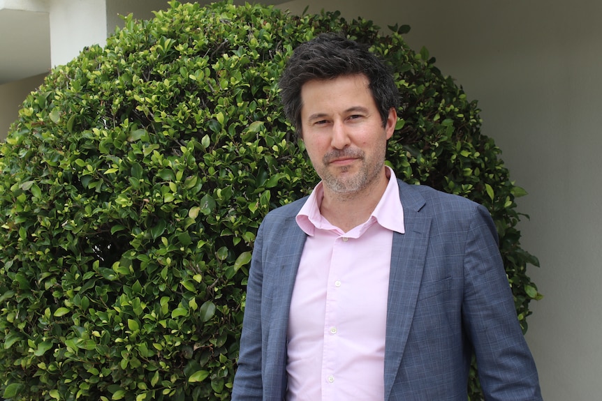 Portrait of Matt Tyler from The Men's Project. He wears a light pink shirt and grey blazer and stands in front of a hedge.