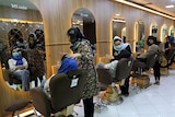 Women in masks and headscarves sit in beauty salon chairs as women work on them. 