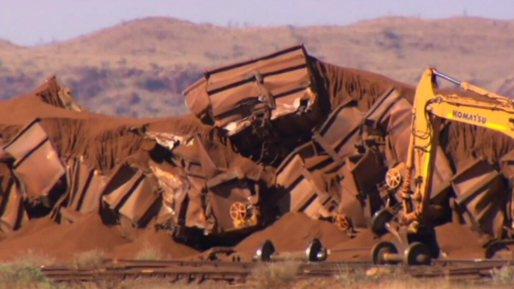 A pile of train wagons lie crumpled on the ground alongside a rail line with axles and a yellow crane nearby.