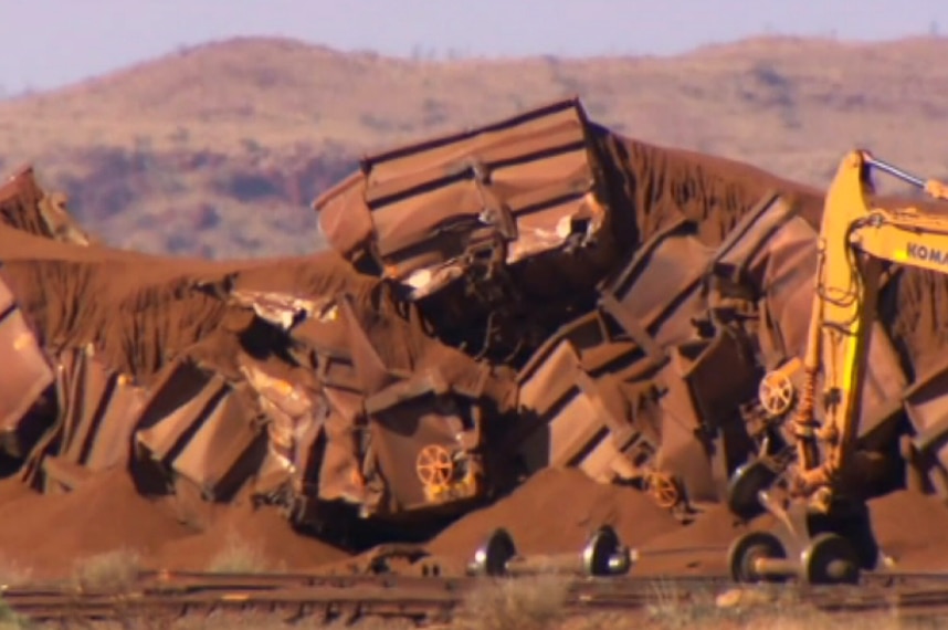 A pile of train wagons lie crumpled on the ground alongside a rail line with axles and a yellow crane nearby.
