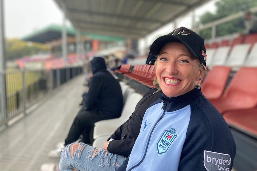 Seorang wanita dengan jaket NSW Blues tersenyum saat dia duduk di tribun kosong.