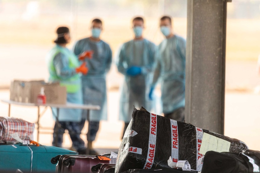 Passengers from India wearing masks and gowns collect their luggage