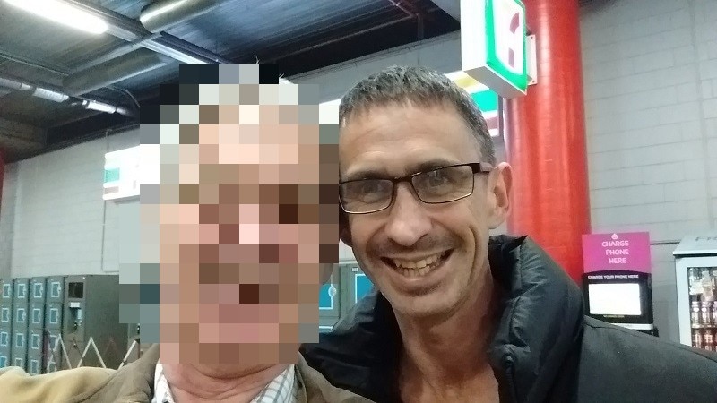 Man with glasses smiles for the camera - the face of the person next to him is pixelated.