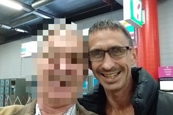Man with glasses smiles for the camera - the face of the person next to him is pixelated.