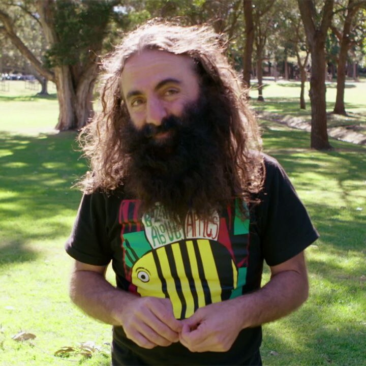 man with long brown hair and beard smiling into camera, he is wearing a bee shirt