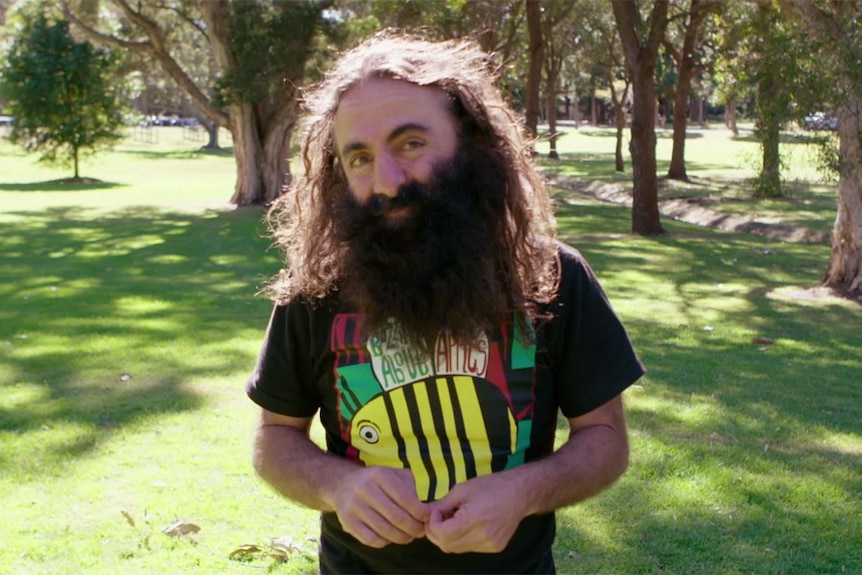 man with long brown hair and beard smiling into camera, he is wearing a bee shirt