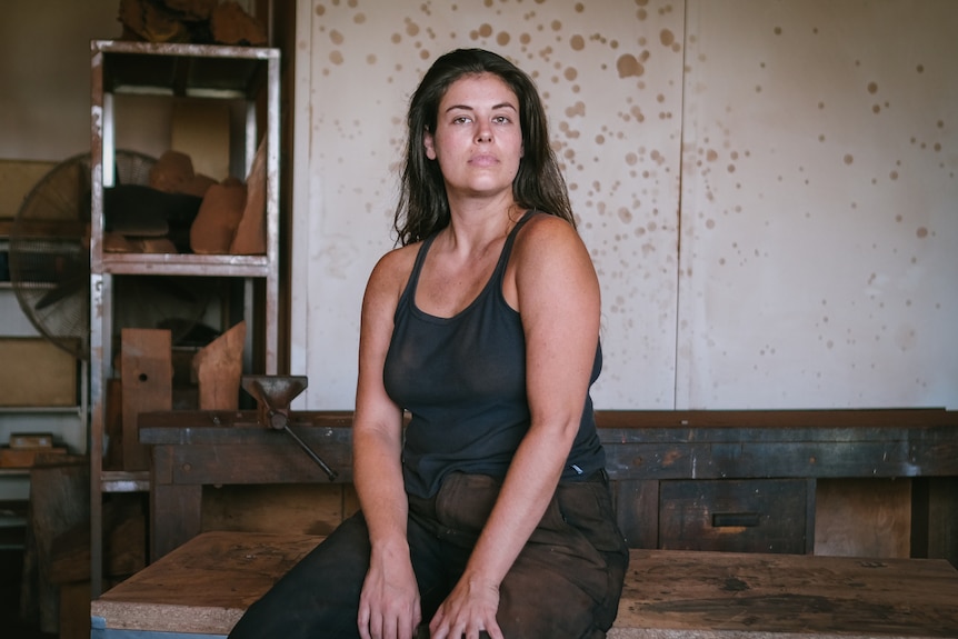 Portrait of artist Olive Gill-Hille, wearing black, sitting on a bench in her studio facing the camera 