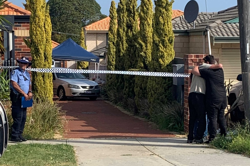 Three people stand hugging next to the driveway of a house with police tape across it.